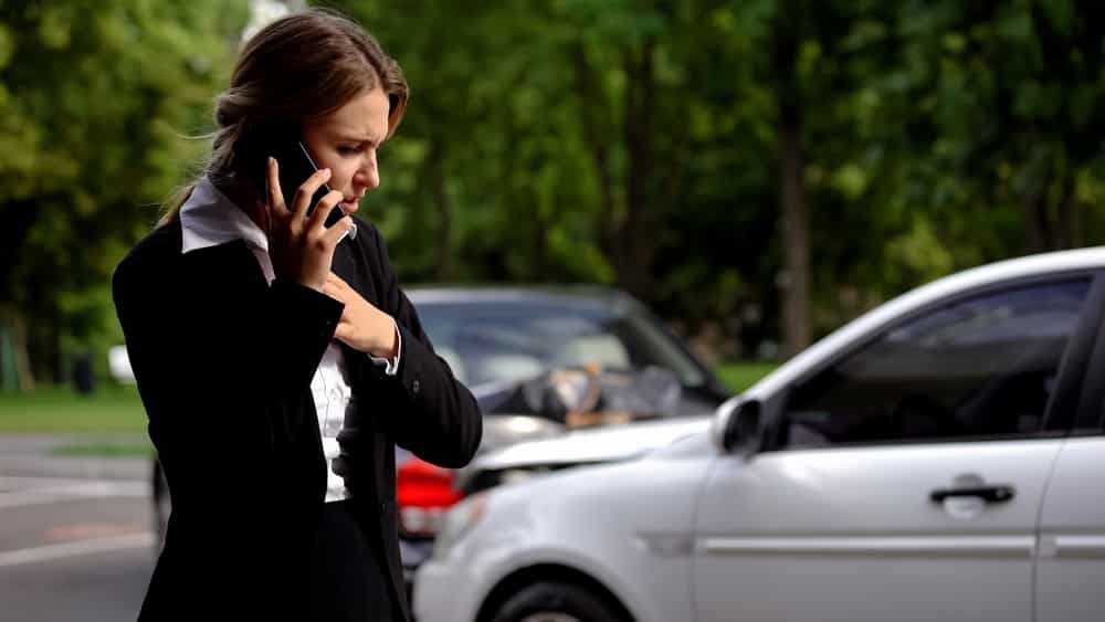 Distressed woman in business attire on phone near damaged car after motor vehicle accident looking worried in outdoor setting
